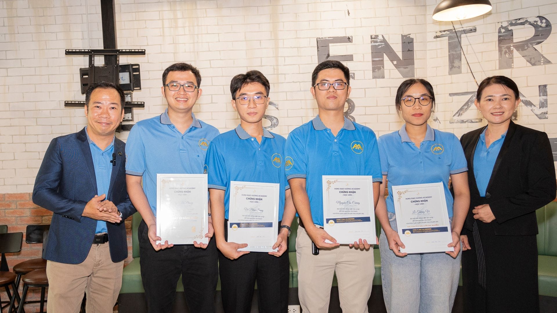 Civil Engineering Outsourcing Six people are gathered indoors, smiling for the camera. Four individuals dressed in matching blue shirts proudly display certificates from the ATED Seminar, while those on either end wear formal business attire. In the background, a white brick wall with partial text hints at a professional environment—reflecting the collaborative spirit often seen in civil engineering outsourcing teams.