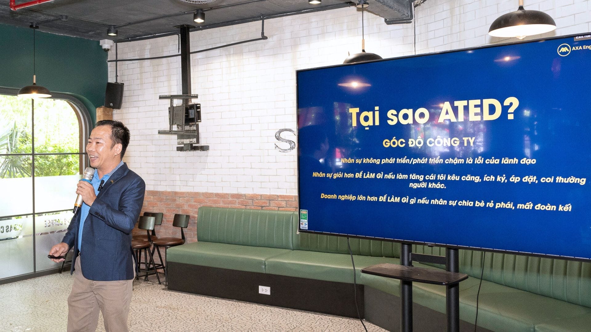 Civil Engineering Outsourcing A man in a blue suit is speaking into a microphone and pointing at a large screen displaying a Vietnamese presentation titled Tại sao ATED? during an ATED seminar centered on civil engineering outsourcing and holistic development, held in a modern, well-lit room with white brick walls.