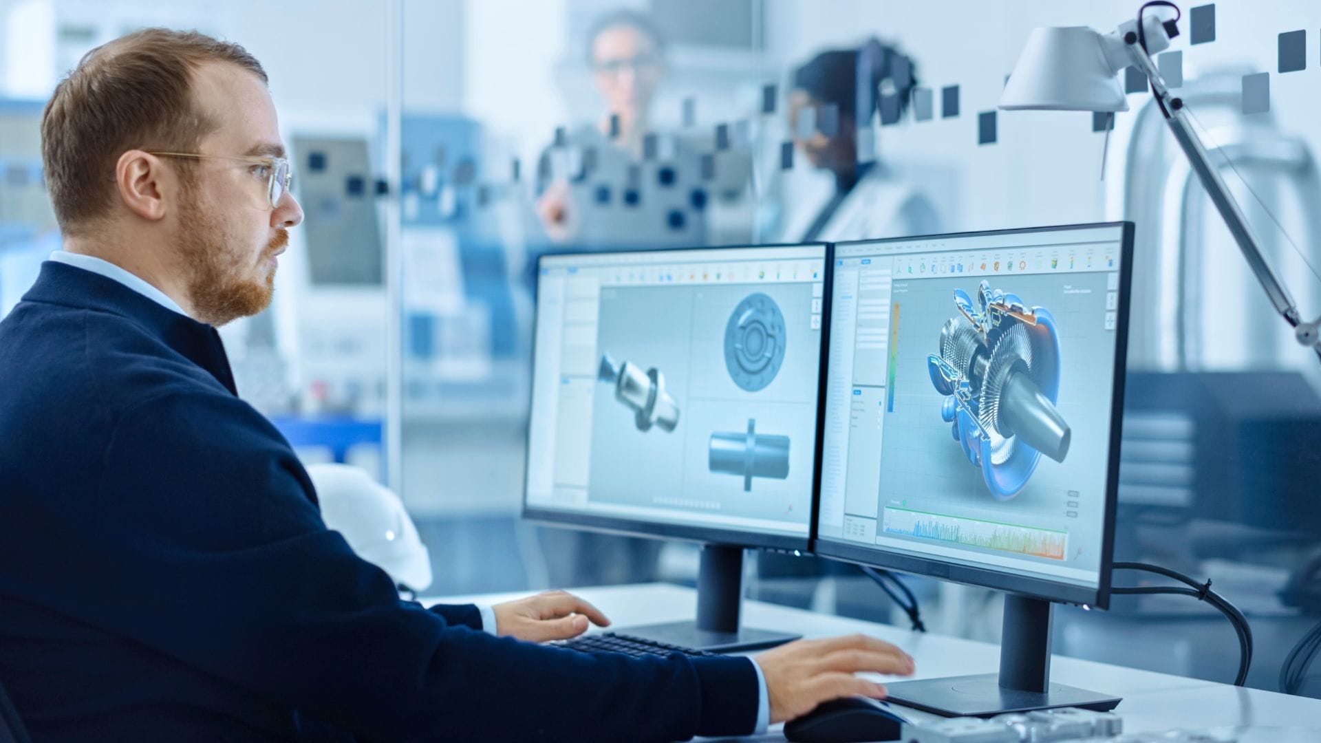 A man with a beard is working on a computer in an office, designing a mechanical component under tight deadlines. He is focused on dual monitors displaying 3D models. Other people are blurred in the background, suggesting a collaborative environment.