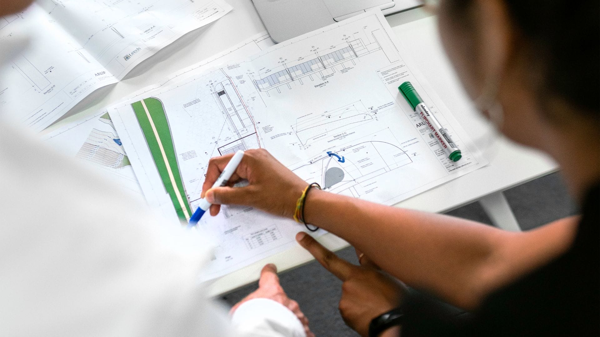 Civil Engineering Outsourcing Two individuals examine technical drawings and blueprints related to civil engineering outsourcing projects spread across a desk. One person highlights specific sections on the documents, which display detailed diagrams and measurements, using a pen; a green marker rests nearby. Risks Associated with Outsourcing Civil Engineering