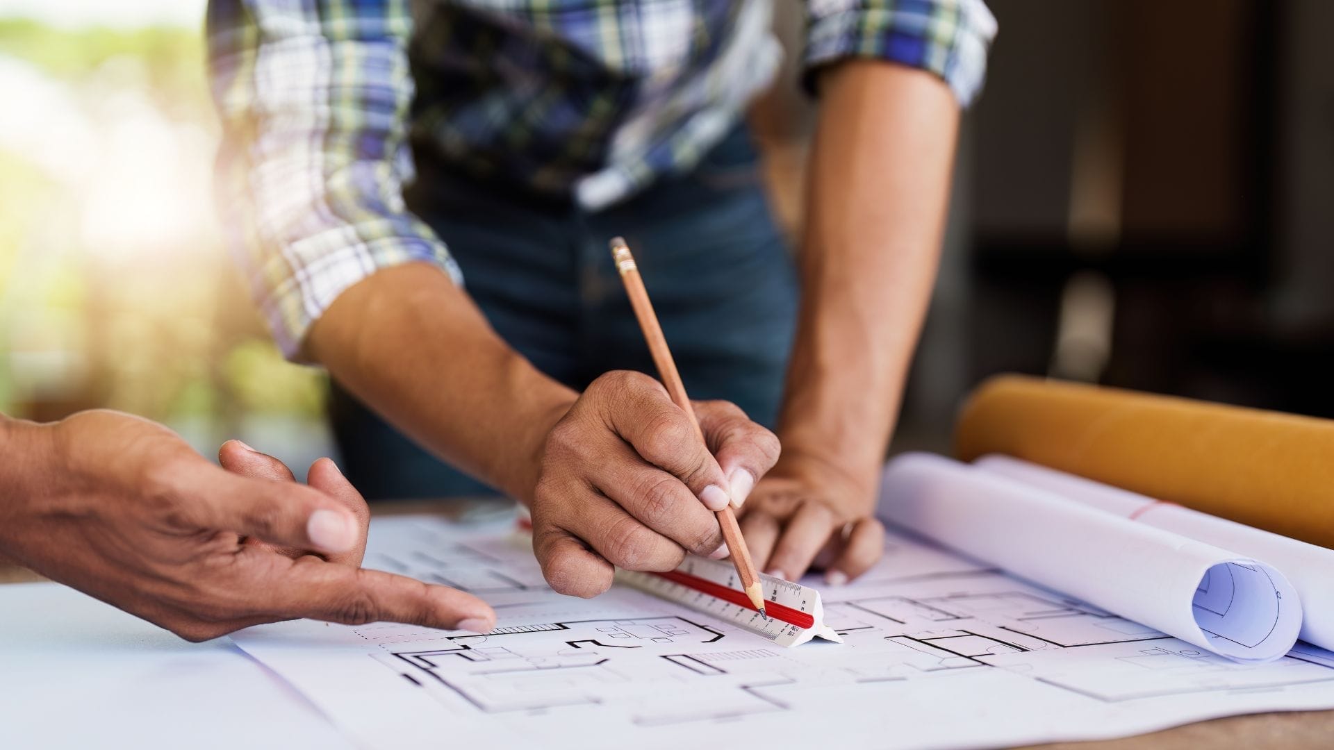 Civil Engineering Outsourcing Two professionals collaborate over architectural blueprints, exemplifying the value of civil engineering outsourcing—with one meticulously drawing precise lines using a pencil and ruler, while the other points out key details on the plans. Rolled-up documents and a sunlit backdrop highlight their expanding capabilities through partnership.