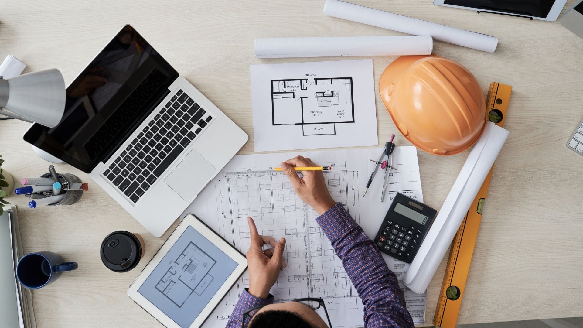 How Outsourcing Is Expanding Capabilities in Civil Engineering Companies 5 Civil Engineering Outsourcing A professional is seated at a desk reviewing architectural blueprints, equipped with a laptop, tablet, calculator, hard hat, ruler, pencil, and coffee cup—demonstrating how civil engineering outsourcing supports the review and enhancement of floor plans while broadening access to modern design to expanding capabilities