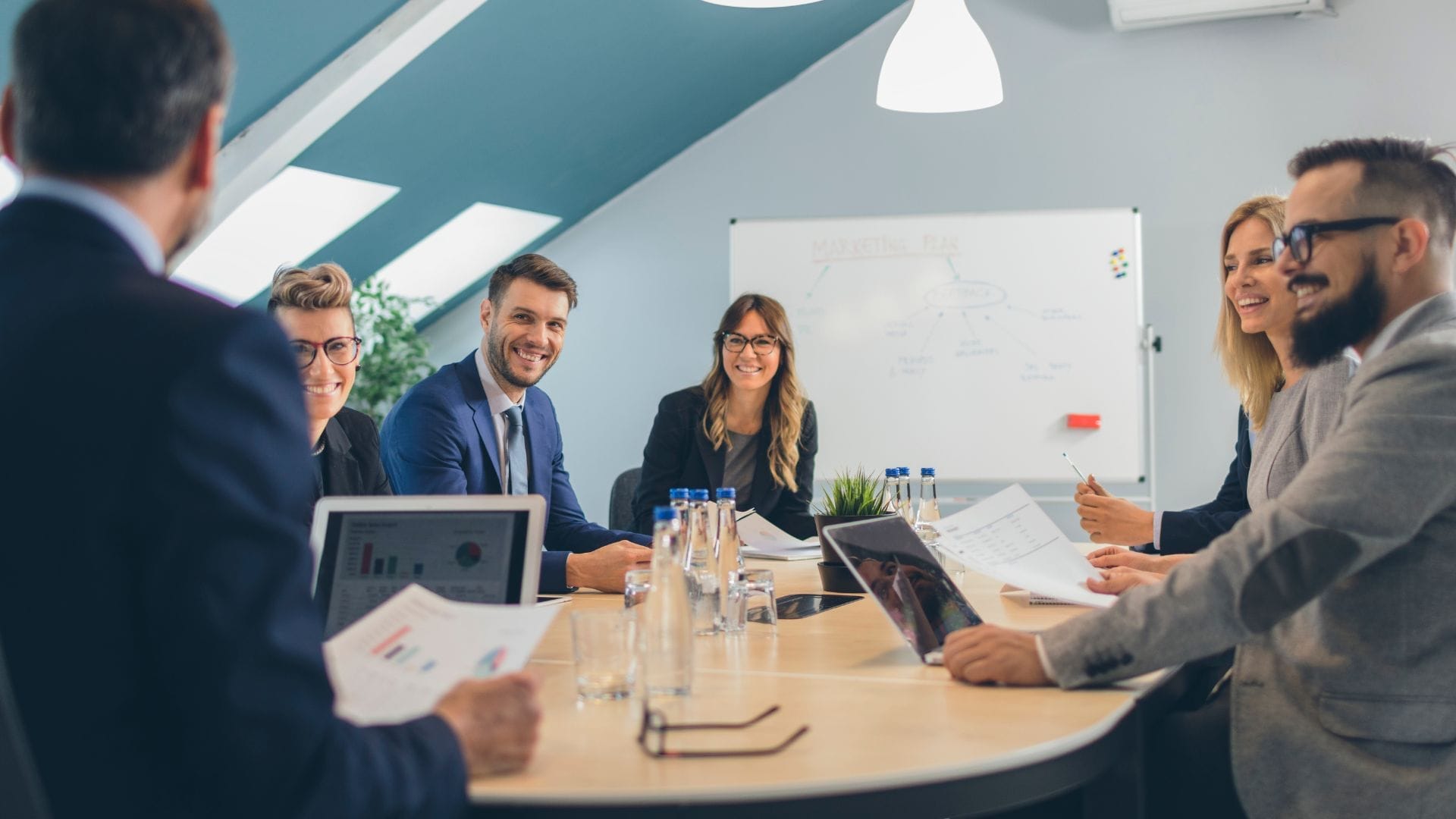 Civil Engineering Outsourcing A team of business professionals is gathered around a conference table, engaged in a positive discussion about civil engineering outsourcing as they review important documents. In the background, a whiteboard displays technical diagrams, while laptops and water bottles are arranged on the table.