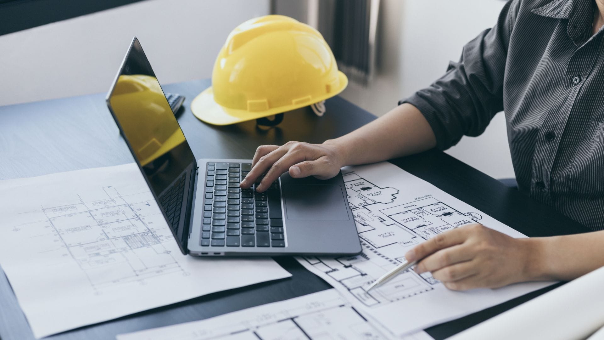 What Are the Trends in Civil Engineering Outsourcing? 7 Civil Engineering Outsourcing An individual works on a laptop at a desk surrounded by architectural blueprints and a yellow construction helmet, illustrating their role in engineering or construction planning and showcasing the prevalence of civil engineering outsourcing among contemporary industry trends.