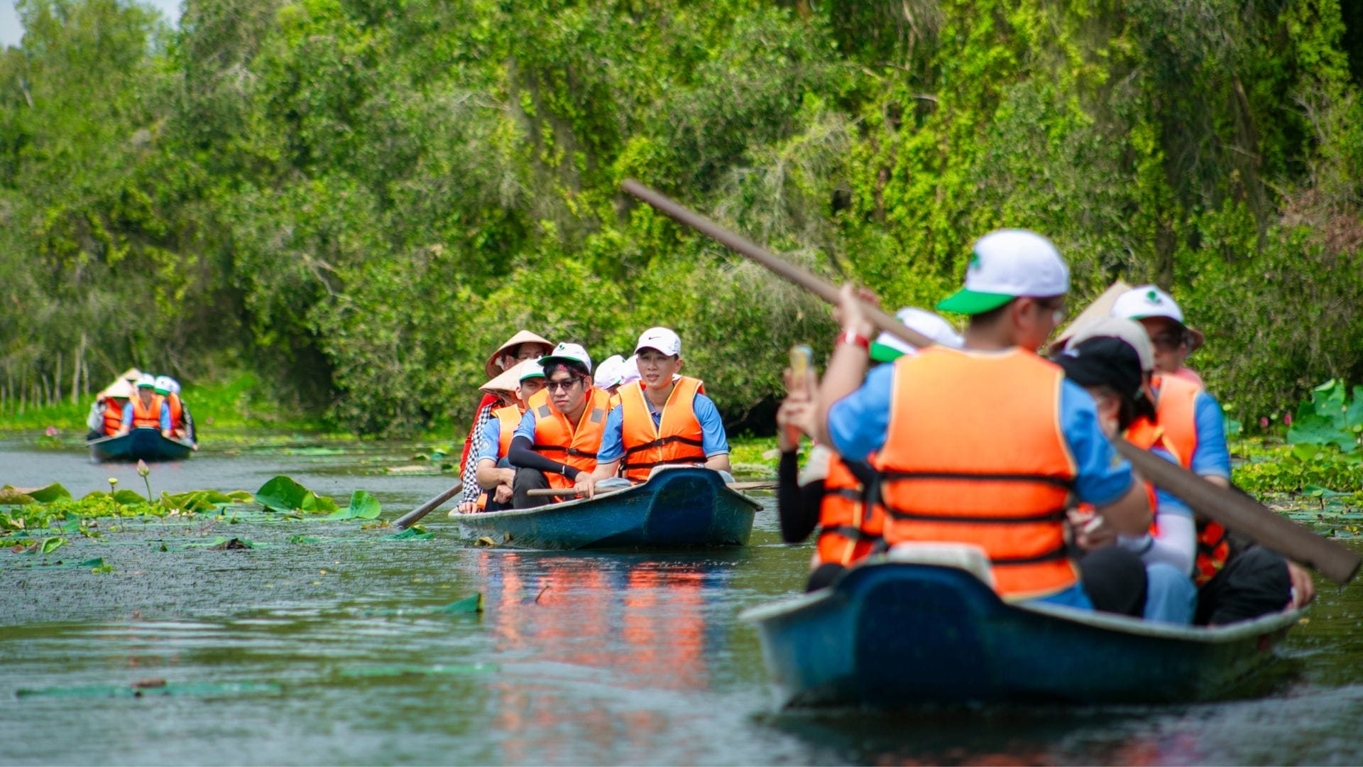 Civil Engineering Outsourcing A group of people wearing orange life jackets and hats paddle canoes along a lush, green waterway surrounded by dense vegetation—creating meaningful moments on the KELTIC–AXA Engineers Trip 2025, which highlights the positive impact of civil engineering outsourcing in enhancing natural environments and recreational experiences.