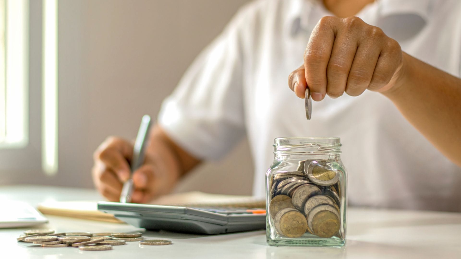 Improve Quality Of Civil Engineering Projects With Outsourcing 7 Civil Engineering Outsourcing A person drops coins into a glass jar filled with coins while jotting down notes and using a calculator, illustrating how civil engineering outsourcing can lead to cost savings, improved financial planning, and enhanced project quality.