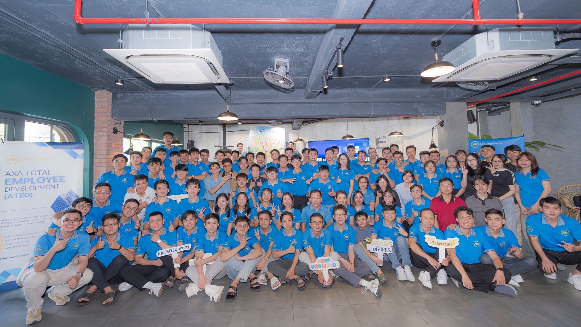 Civil Engineering Outsourcing A large group in matching blue shirts pose together indoors at a company event, smiling for the camera. Some hold signs with positive messages about holistic growth, celebrating their achievements in civil engineering outsourcing. Banners and a modern ceiling set the scene as they embark on their ATED 2025 journey.