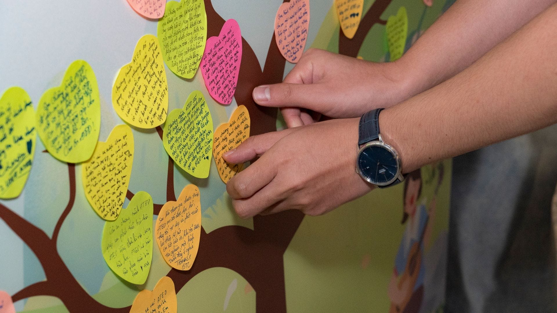 Civil Engineering Outsourcing A person wearing a watch attaches a yellow, heart-shaped note with handwritten text to a wall display resembling a tree filled with similarly colorful heart-shaped notes. This vibrant display symbolizes their journey toward holistic growth in the field of civil engineering outsourcing at ATED 2025.