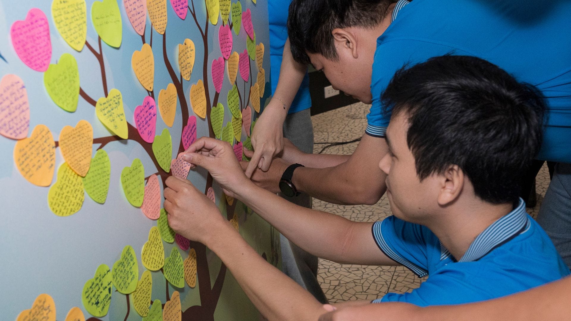 Civil Engineering Outsourcing Two individuals in blue shirts are affixing vibrant, heart-shaped notes with handwritten messages to a wall featuring a tree illustration—representing their comprehensive growth experience through civil engineering outsourcing. Additional notes, already attached to the tree’s branches, further highlight collective progress and shared insights.