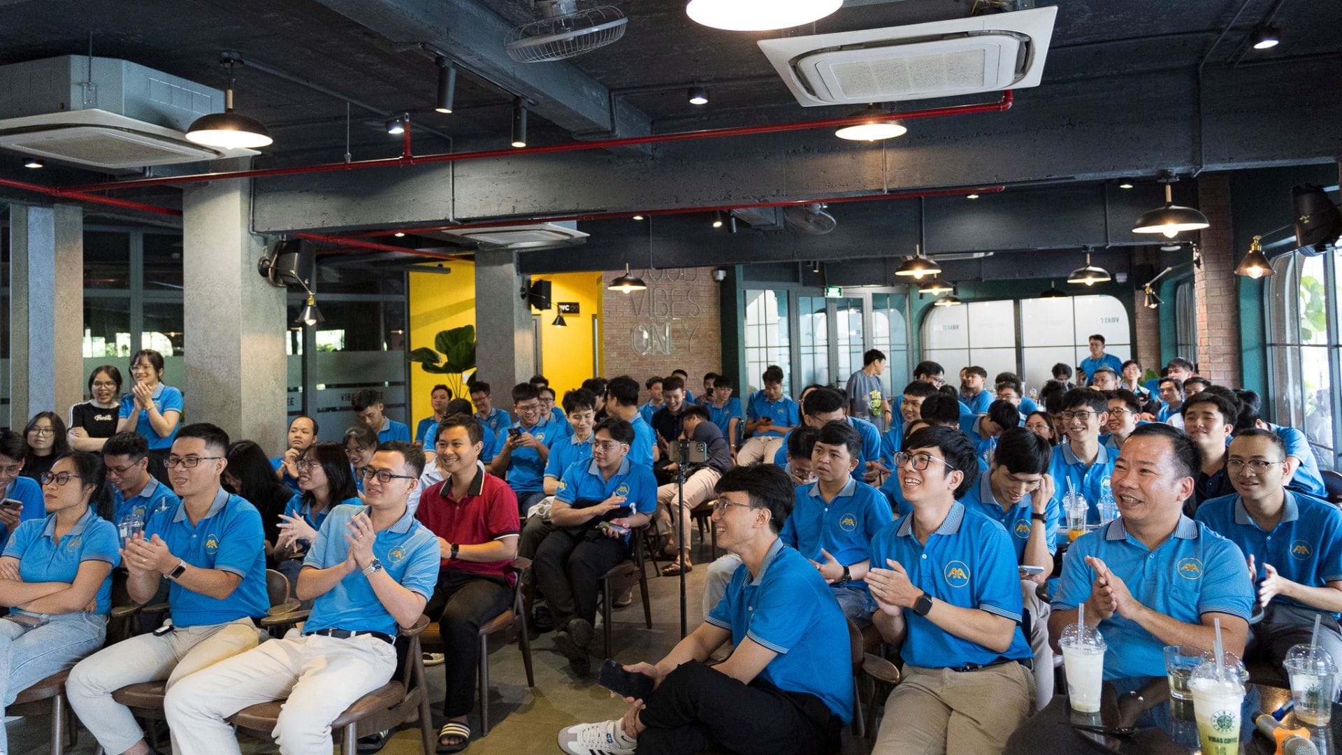 Civil Engineering Outsourcing A large group in matching blue shirts gathers in a modern, industrial-style room, smiling and applauding at an indoor ATED 2025 event focused on civil engineering outsourcing. With drinks set on tables and a vibrant atmosphere all around, the attendees reflect a shared commitment to holistic growth through collaboration within the field.