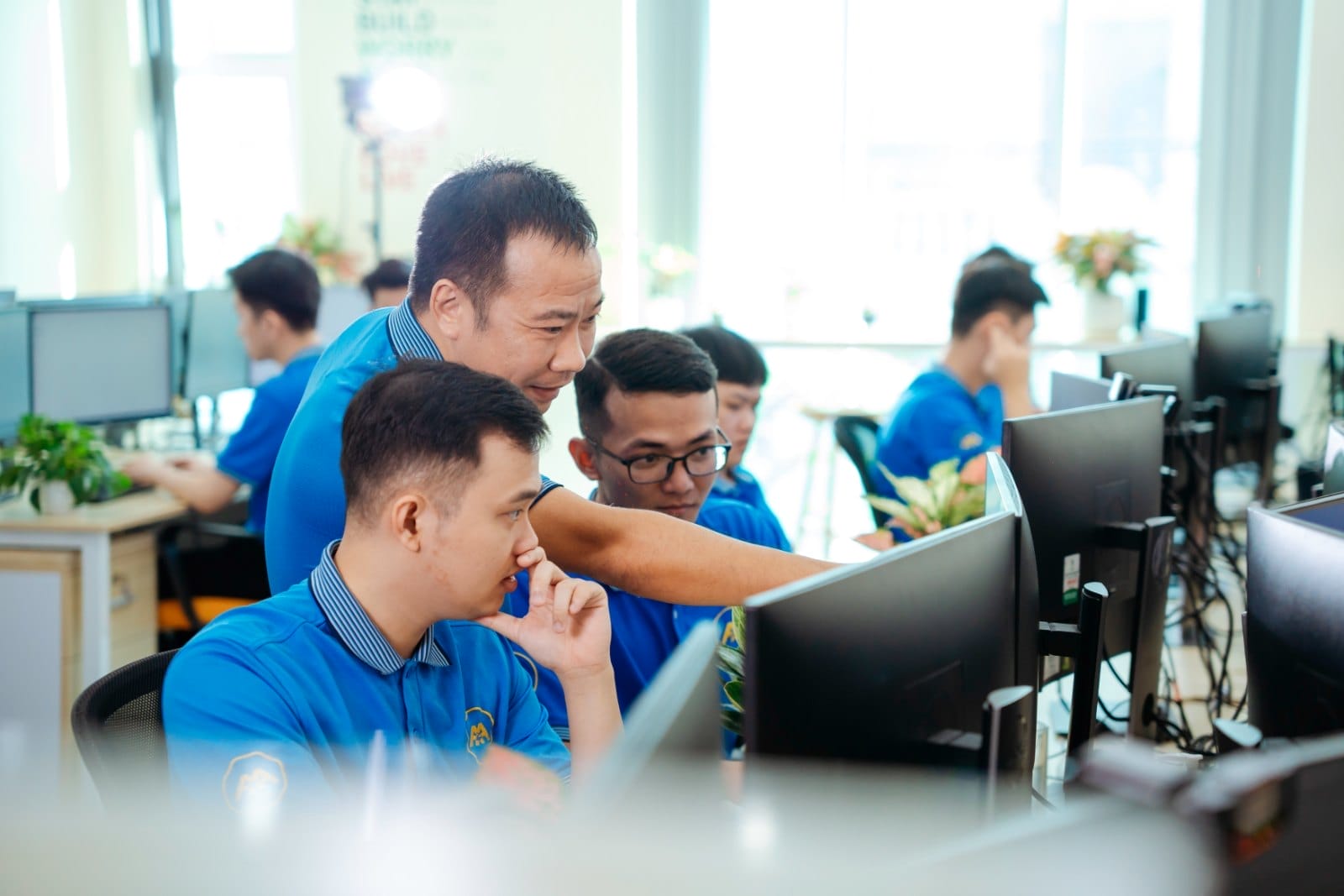 Civil 3D 2026: Innovation That Actually Works for Real-World Engineering 6 Civil Engineering Outsourcing A team of civil engineering outsourcing professionals in blue shirts collaborate at computer desks in a well-lit office. One team member stands, pointing at a monitor as colleagues listen attentively. The workstations feature computer screens and plants, creating an organized and productive environment.