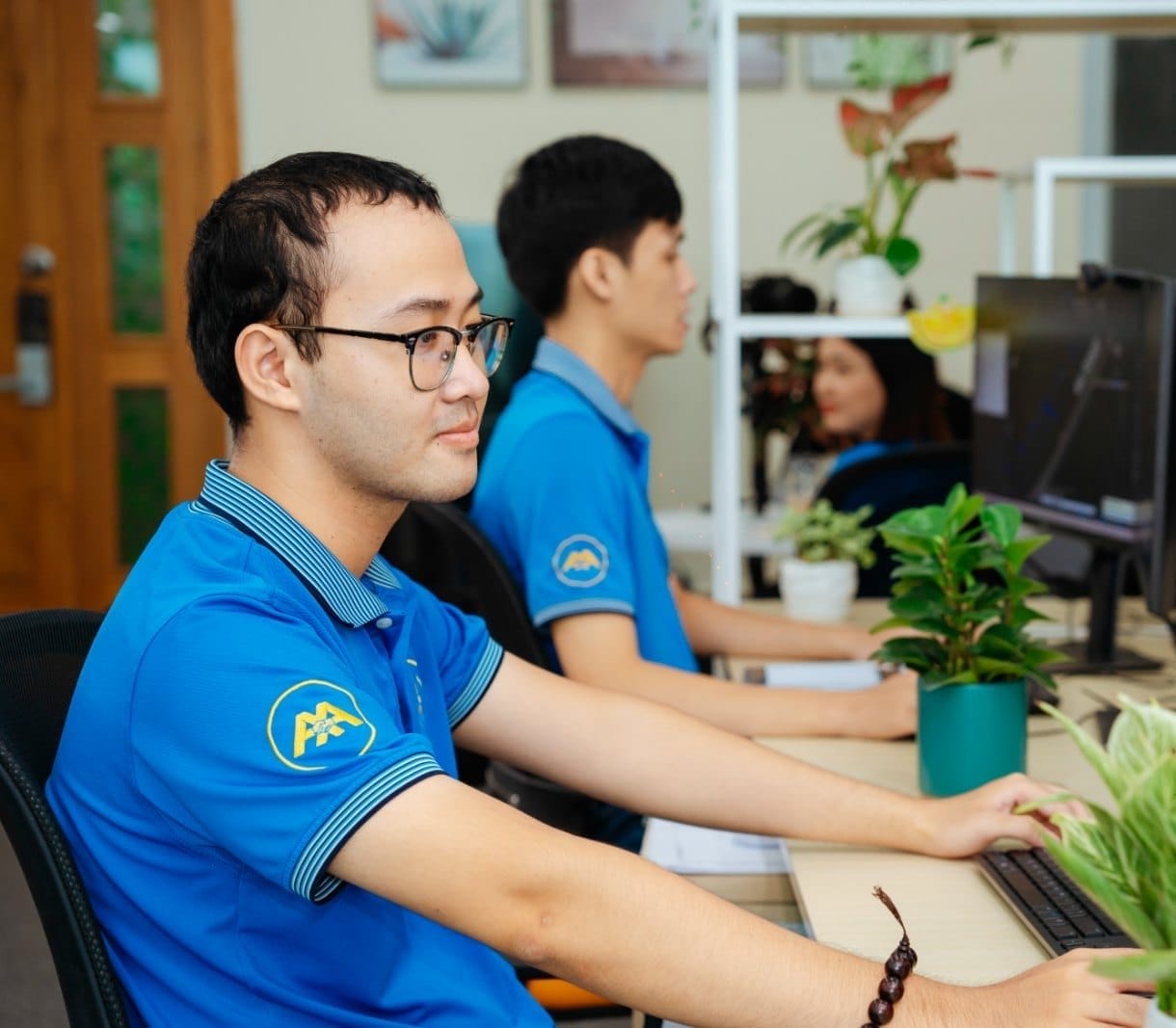 Civil Engineering Outsourcing Three professionals in blue shirts work at desks with computers in a modern office specializing in civil engineering outsourcing. Lush green potted plants decorate the desks, while additional greenery is arranged on shelves in the background.