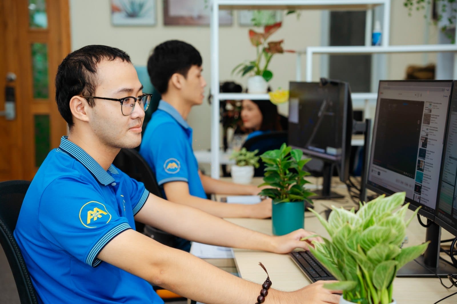 Civil 3D 2026: Innovation That Actually Works for Real-World Engineering 5 Civil Engineering Outsourcing Three professionals in blue shirts work at desks with computers in a modern office specializing in civil engineering outsourcing. Lush green potted plants decorate the desks, while additional greenery is arranged on shelves in the background.