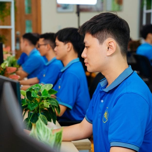 Civil Engineering Outsourcing Young men in blue shirts work at computers in a civil engineering outsourcing office, focused amid potted plants and a collaborative vibe.