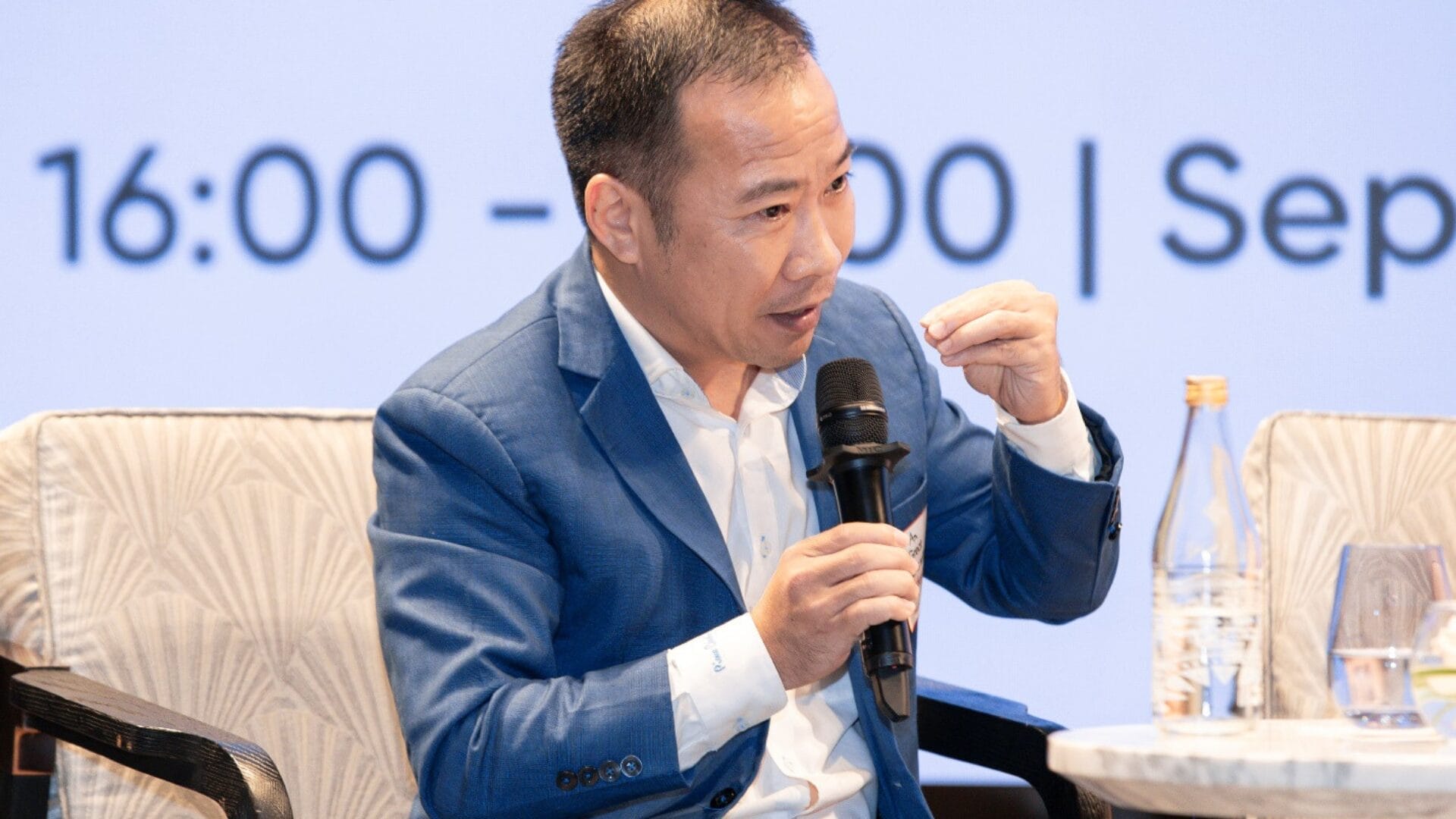 Workplace Culture Masterclass 04 — The Journey of Nurturing Culture at AXA Engineers 6 Civil Engineering Outsourcing A man in a blue suit speaks at an AXA Engineers panel on civil engineering outsourcing, gesturing beside a water bottle and glass.