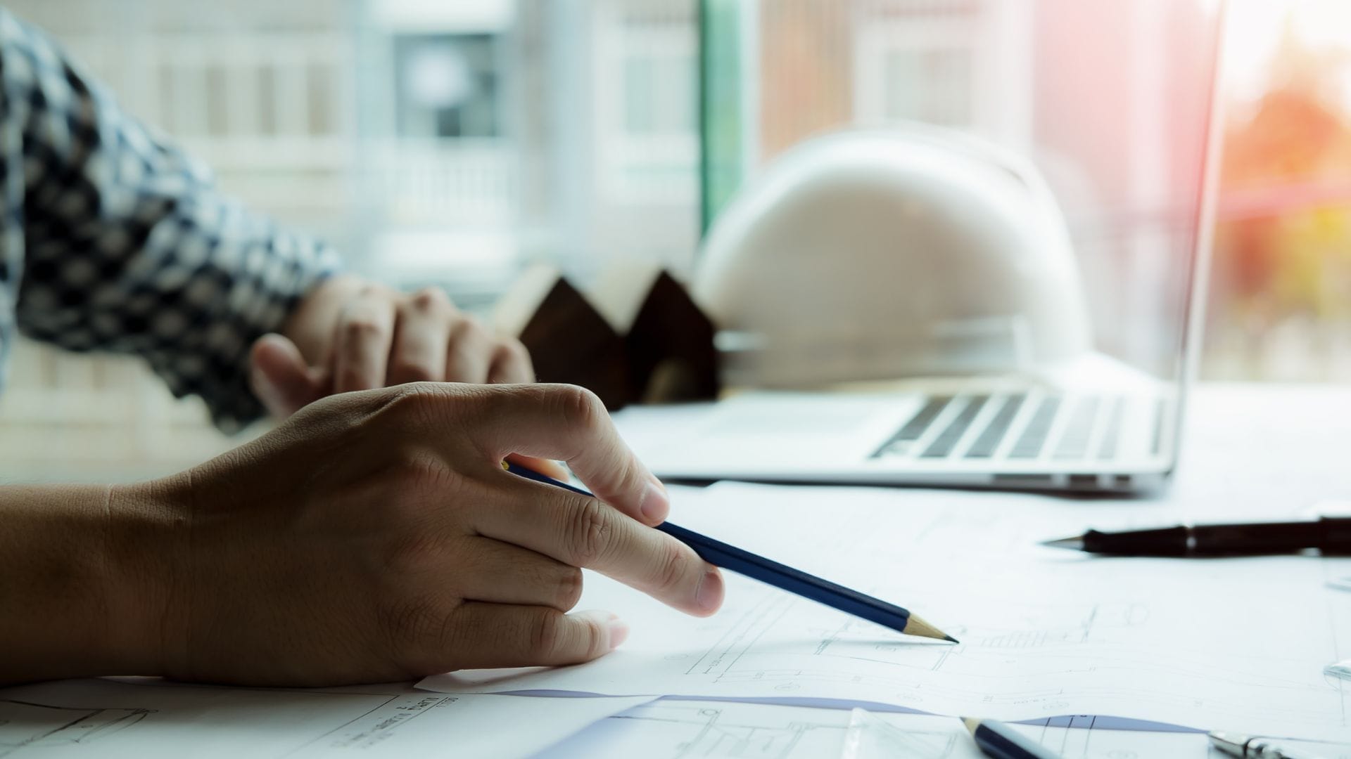 Civil Engineering Outsourcing A person reviews blueprints at a desk with a laptop, hard hat, and model house, highlighting civil engineering outsourcing today.