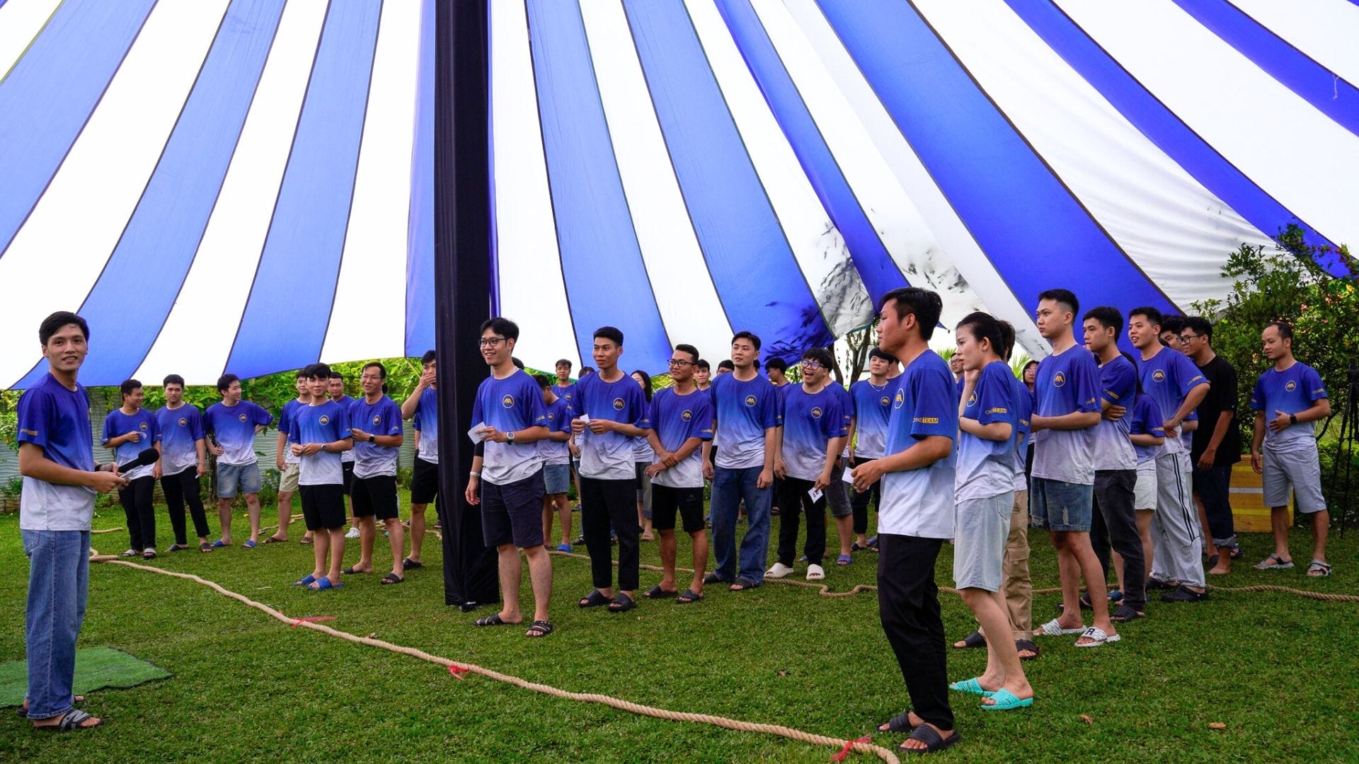 Civil Engineering Outsourcing A group in matching blue and white shirts stands on grass under a tent, listening to an AXA Engineers leader discuss civil engineering outsourcing.
