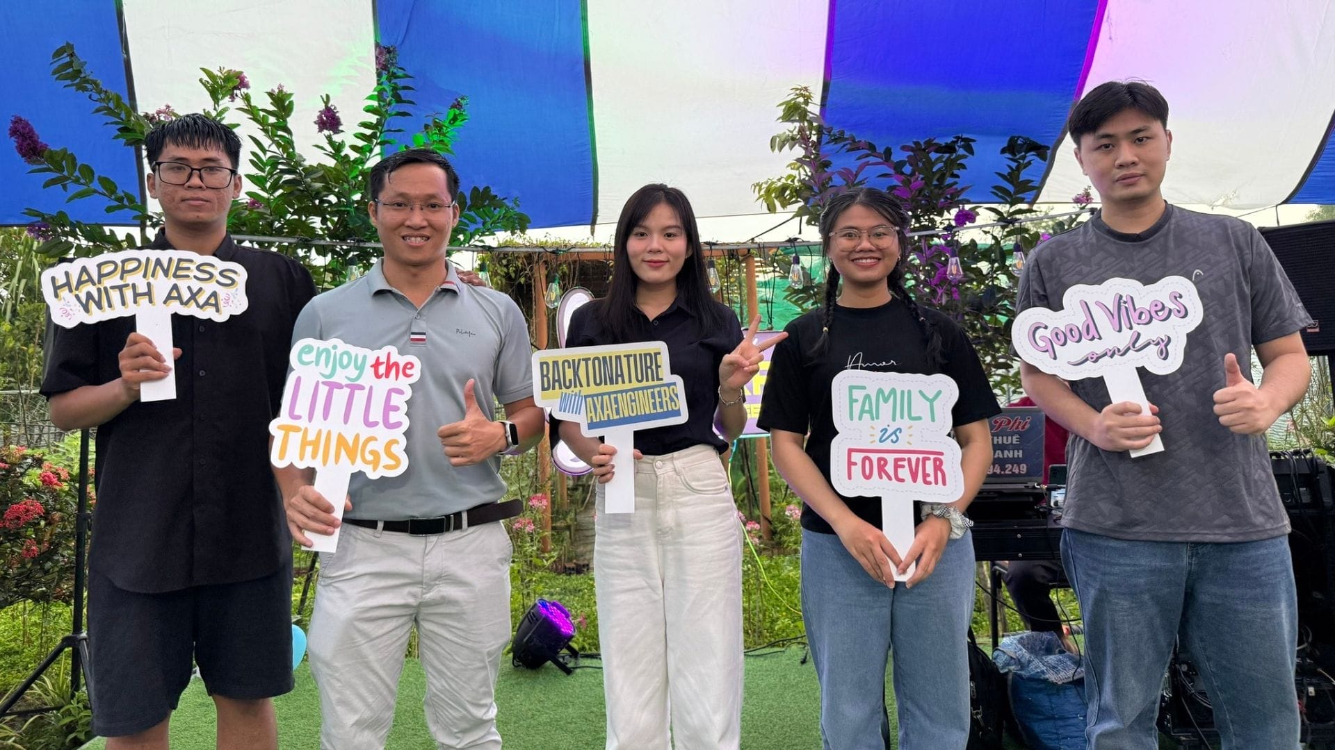 Civil Engineering Outsourcing Five people at the AXA Engineers Garden tent, holding positive signs about civil engineering outsourcing, smiling with greenery behind.