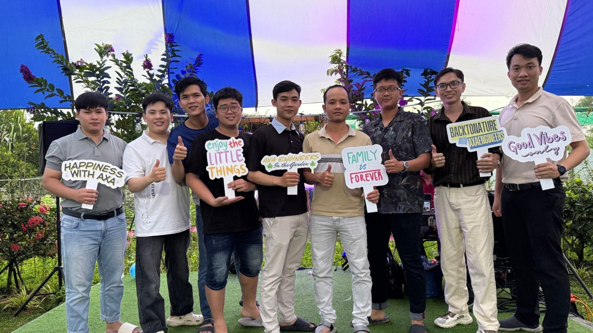 Civil Engineering Outsourcing Nine men stand under a blue-and-white canopy at Engineers Garden, holding signs promoting civil engineering outsourcing and smiling.