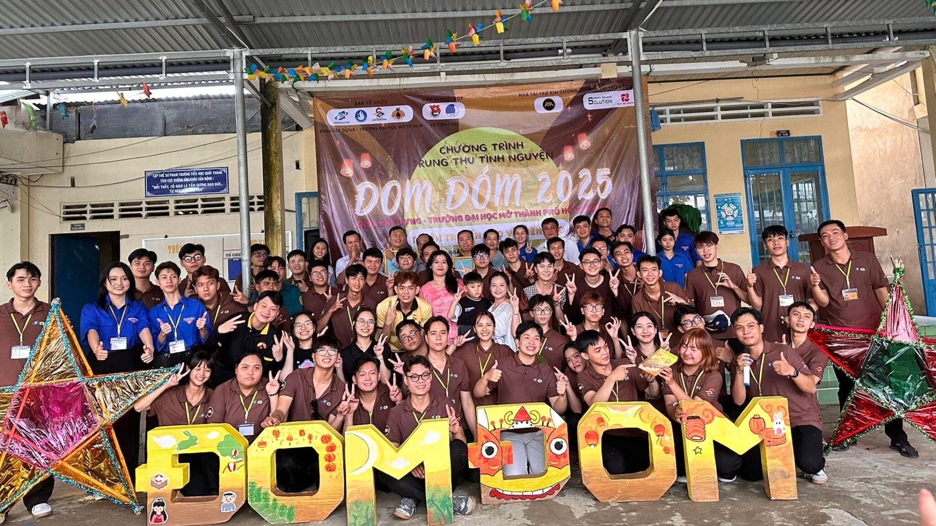 Mid-Autumn Festival Night – The Light of Love 7 Civil Engineering Outsourcing A group of young people, including civil engineering outsourcing volunteers, smile by a Journey of Fireflies 2025 banner with signs and lanterns.