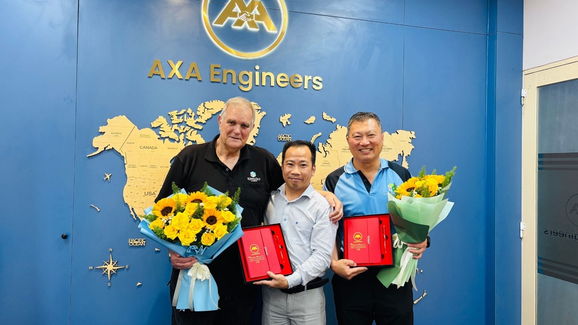 Civil Engineering Outsourcing Three men celebrate civil engineering outsourcing in front of the AXA Engineers logo, holding flowers and plaques, smiling together.