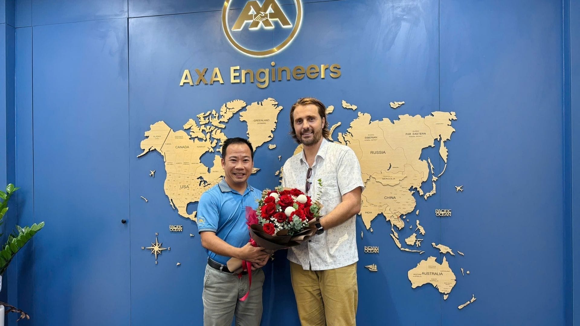 Civil Engineering Outsourcing Two men smile in front of a blue world map wall with “AXA Engineers”; civil engineering outsourcing partners celebrate an occasion.