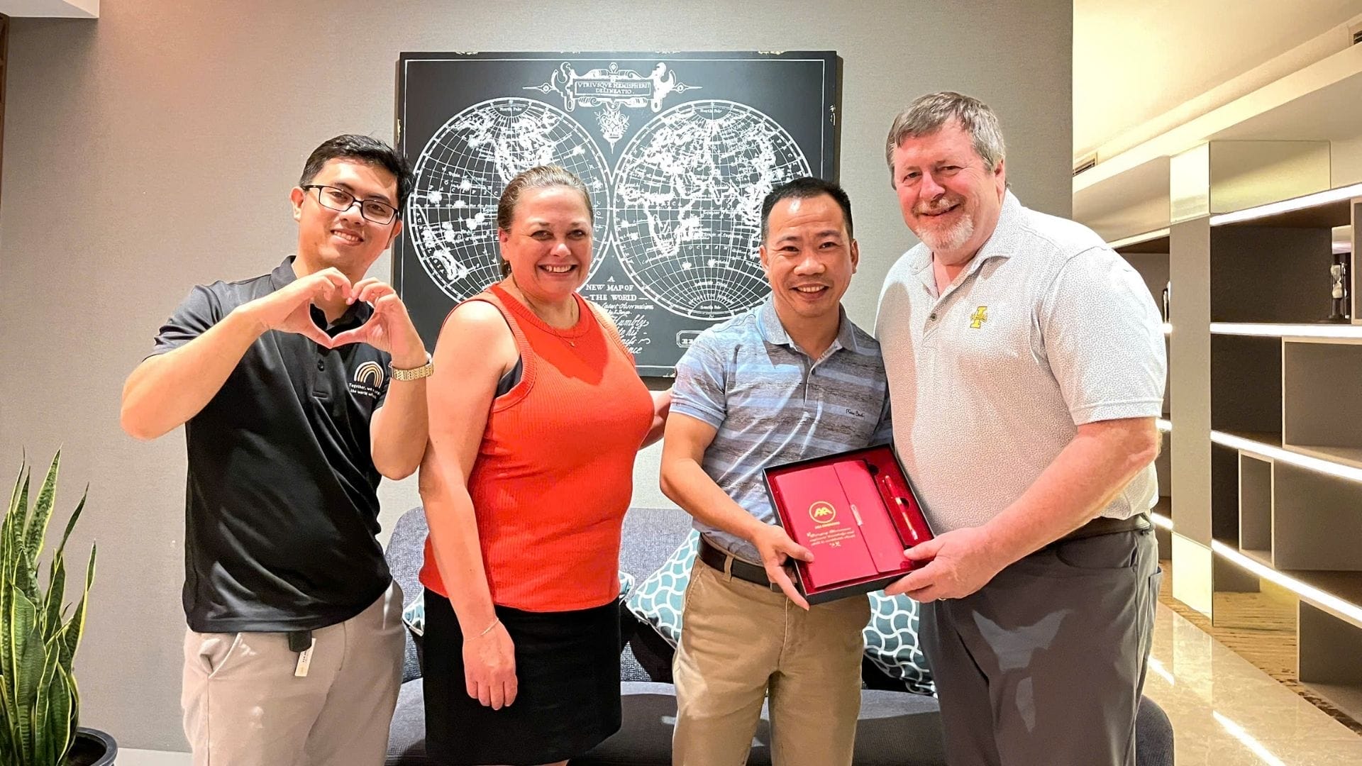 Civil Engineering Outsourcing Four people smile indoors celebrating a civil engineering outsourcing visit; one holds a red box, another makes a heart gesture.