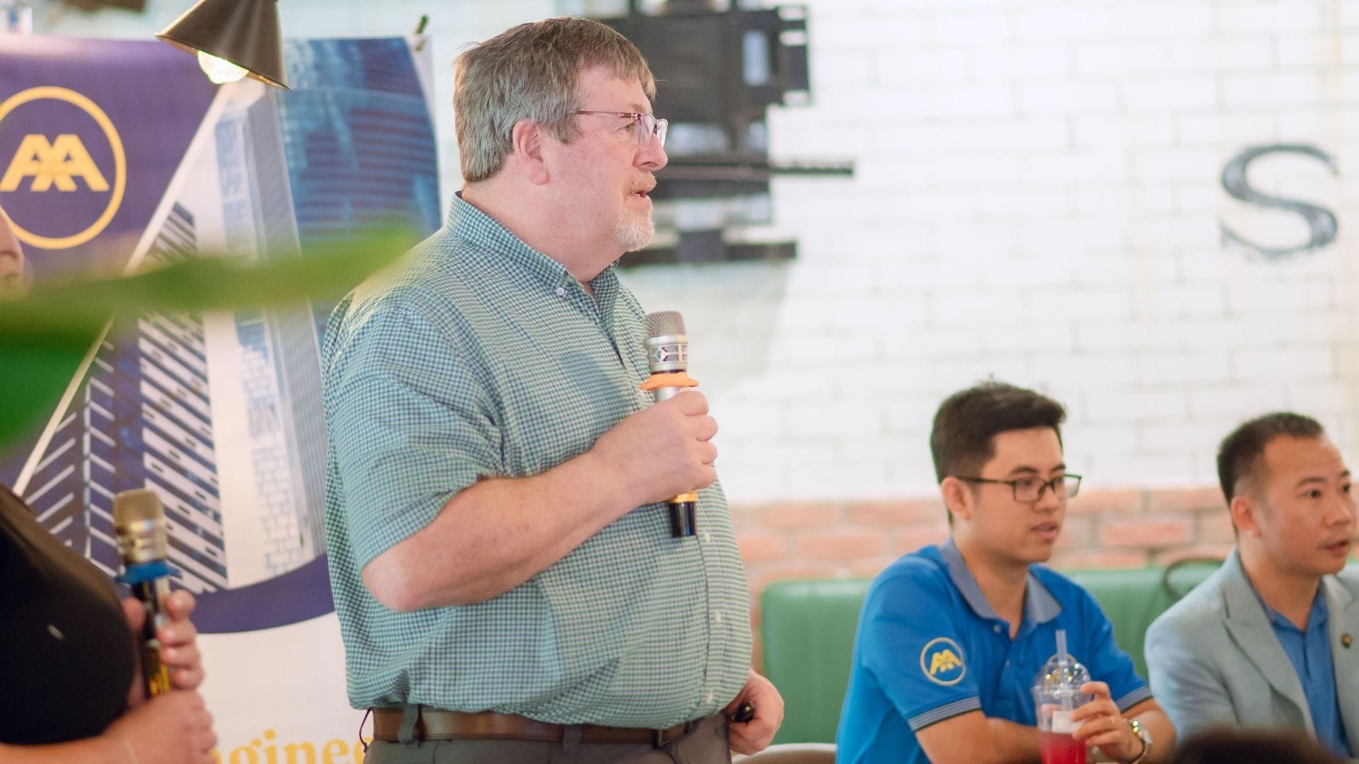 Civil Engineering Outsourcing A man in a green checkered shirt speaks into a microphone at a civil engineering outsourcing event, with two men seated beside him.
