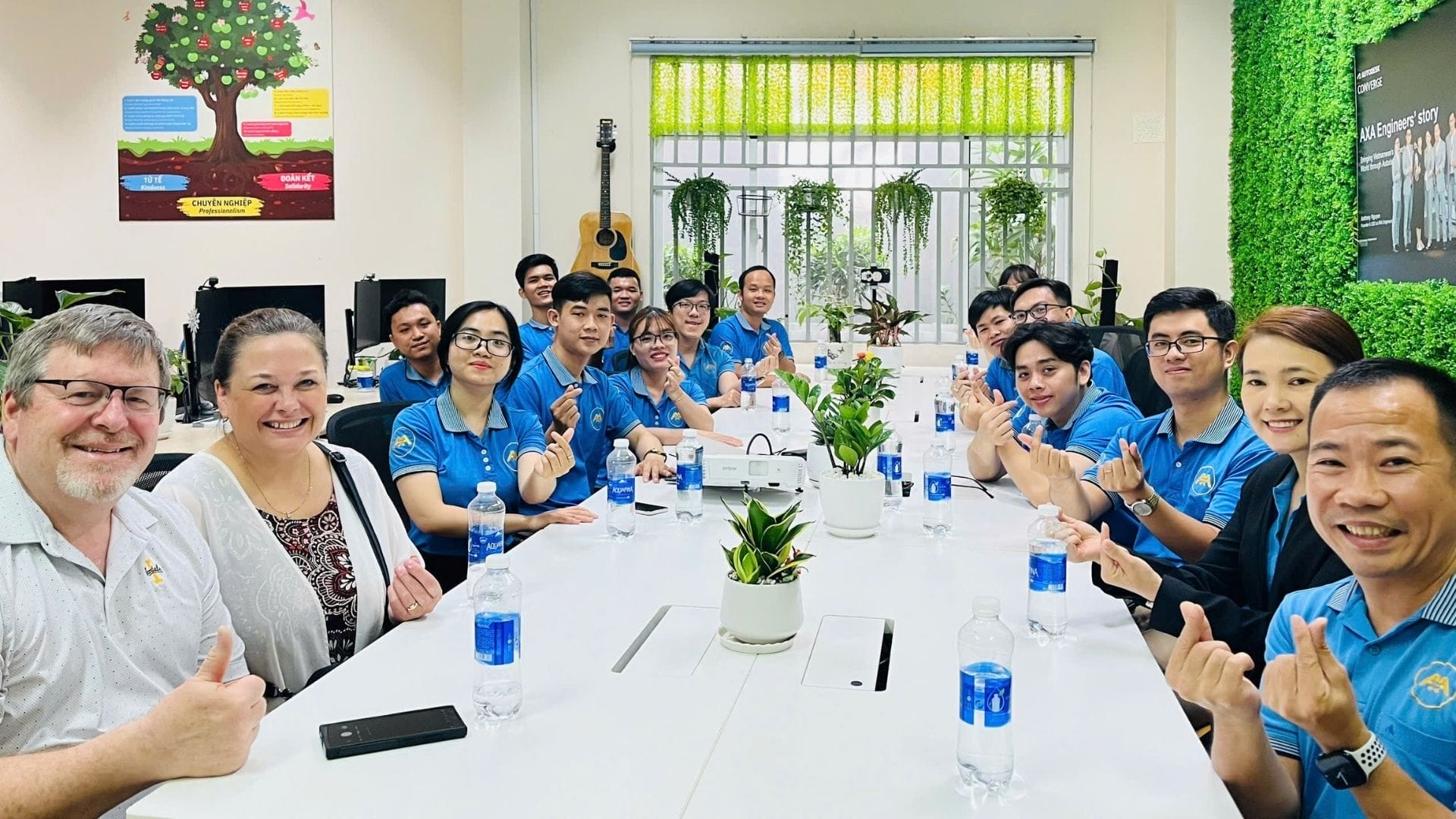 Civil Engineering Outsourcing A team, mainly in blue uniforms, gathers at a white table for a civil engineering outsourcing meeting, smiling and making hearts.