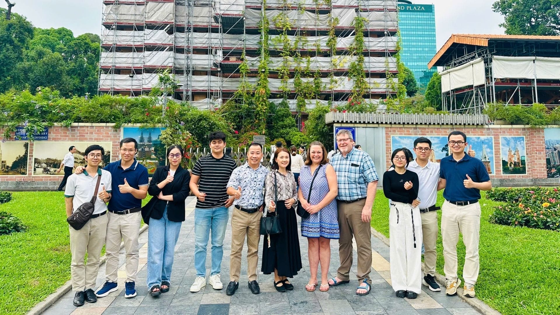Civil Engineering Outsourcing Thirteen people stand in a row, smiling at a civil engineering outsourcing event with greenery, construction, and tall glass building.
